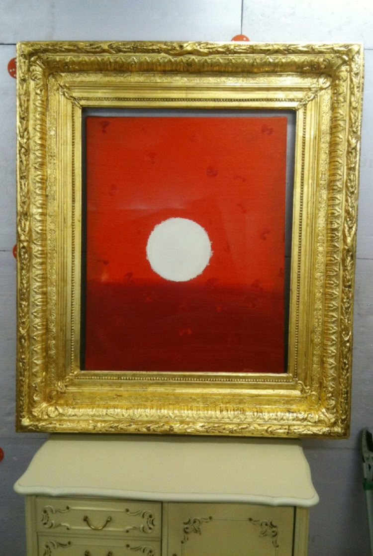 Gemälde mit rotem Hintergrund und weißem Kreis in einem goldenen Rahmen, auf einem hellen Möbelstück stehend.