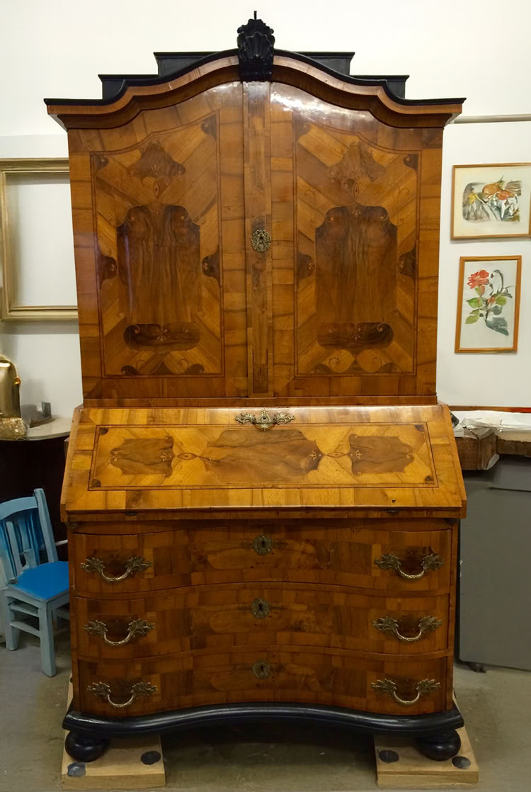 Antiker Holzschrank mit Intarsien, bestehend aus einem oberen Teil mit Türen und einem unteren Teil mit Schubladen, umgeben von gerahmten Bildern und einem blauen Stuhl.