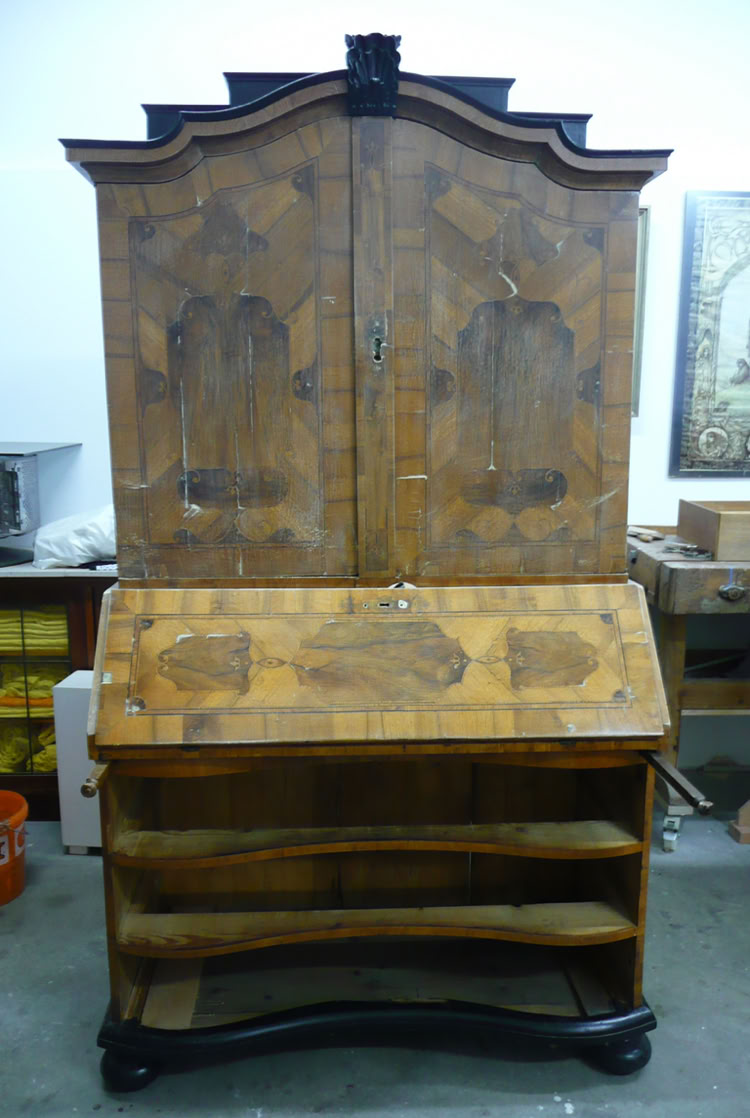 Antiker Holzschrank mit verzierten Türen und einem herunterklappbaren Schreibpult, darunter offene Regalfächer.